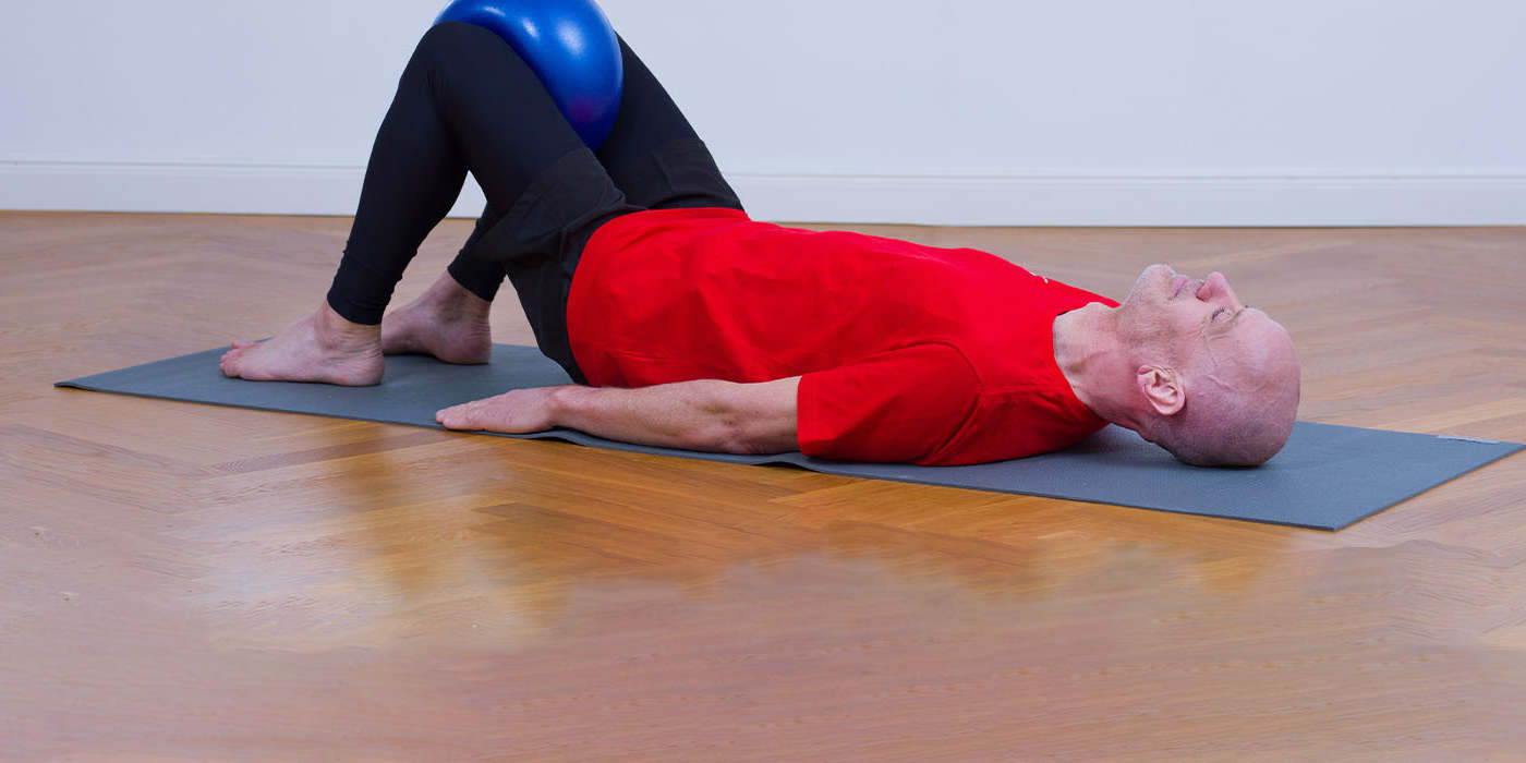 Älterer Mann liegt auf Gymnastikmatte und presst mit seinen Knien einen Ball zusammen Älterer Mann liegt auf Gymnastikmatte und presst mit seinen Knien einen Ball zusammen